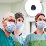 Gros plan sur trois professionnels de la santé, dont un chirurgien portant des lunettes et deux collègues, tous en tenue chirurgicale complète (blouse, masque, calot à motifs) regardant attentivement hors champ dans un bloc opératoire moderne sous un éclairage scialytique. Cette image illustre la collaboration nécessaire lors d'un processus de mobilité professionnelle vers une spécialisation chirurgicale